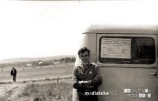 Portret chłopaka na tle samochodu z programem XXIII Ogólnopolskich Zawodów Modeli Latających Spółdzielczości Mieszkaniowej, Lotnisko Krywlany, Białystok, lata 60. XX w.