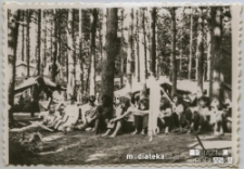 Harcerze siedzący na ziemi na tle obozowiska w lesie