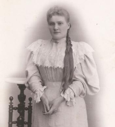 Młoda kobieta z włosami splecionymi w warkocz, suknia w paski, kołnierz i mankiety koronkowe, lewym łokciem oparta o stolik