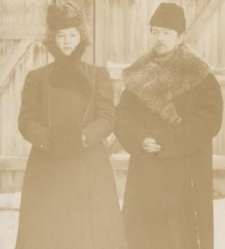 Kobieta w długiej sukni , płaszczu z mufką i kapeluszu, obok mężczyzna w palcie z futrzanym kołnierzemi wysokiej czapce