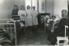 Sala w szpitalu pod ścianami łóżka z pacjentami, stoją trzy pielęgniarki w białych fartuchach i jedna pacjentka w ciemnym szlafroku