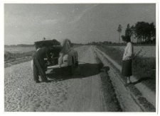 Kobieta stojąca na poboczu drogi. Obok mężczyzna i samochód marki Warszawa