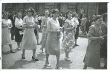 Grupa kobiet idących ulicą, za nimi stoją ludzie na chodniku i budynek